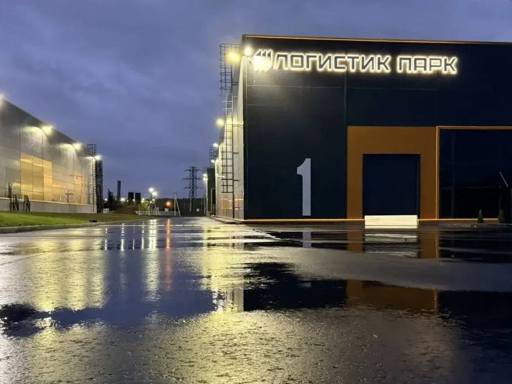 Корпус 1 Логистик Парка Толбино вечером: светящаяся вывеска на тёмном фасаде, жёлтый портал ворот, мокрый асфальт